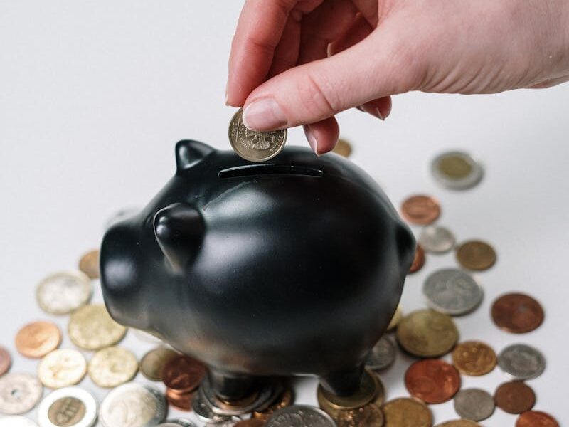Hand inserting coin into a black piggy bank surrounded by various coins, symbolizing savings.